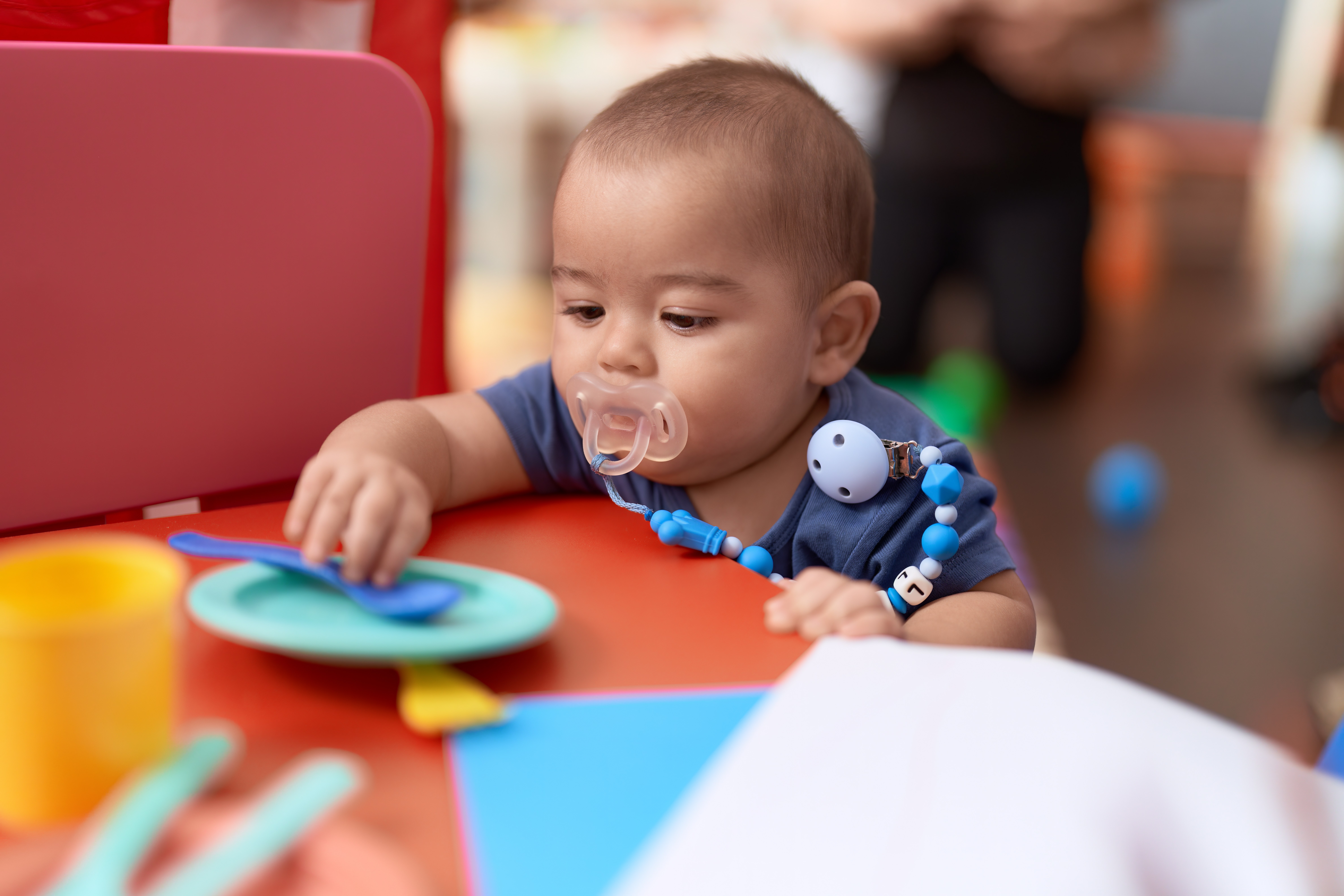 Adorable chinese toddler using pacifier holding dish toy at kindergarten
